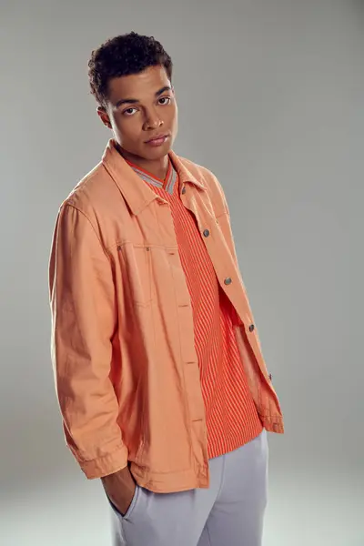 Junger afrikanisch-amerikanischer Mann in pfirsichfarbenem Hemd blickt in die Kamera vor grauem Hintergrund, Hand in Tasche — Stockfoto