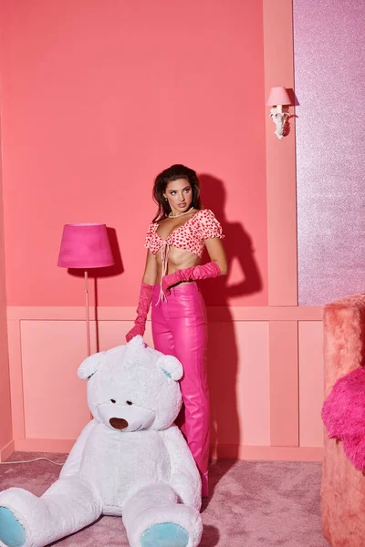 Chic jeune femme en gants, haut de culture rose et pantalon posant près ours en peluche géant dans une chambre vibrante — Photo de stock