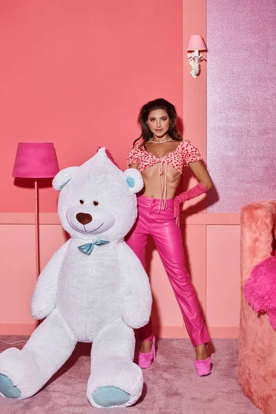 Glamour giovane donna in rosa crop top e pantaloni in posa con mano sul fianco vicino orsacchiotto gigante — Foto stock