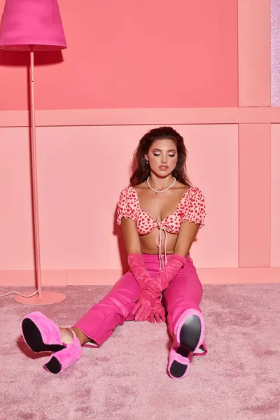 Belle et frisée jeune femme en haut de culture et pantalon assis près du lampadaire dans la chambre rose — Photo de stock