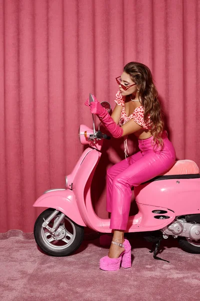 Гламурна молода жінка в стильних сонцезахисних окулярах і сучасному рожевому вбранні сидить на яскравому скутері — стокове фото