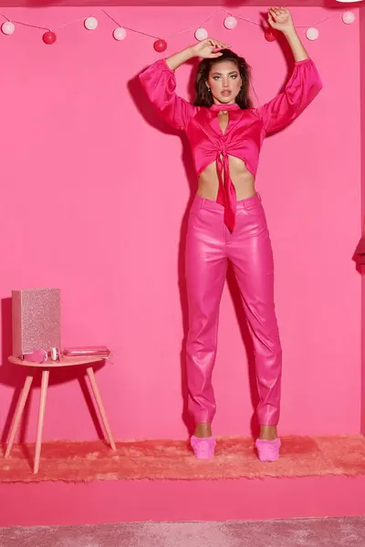 Glamouröse junge Frau in bauchfreiem Top und pinkfarbener Hose, die mit erhobenen Händen neben Ballgirlanden steht — Stockfoto