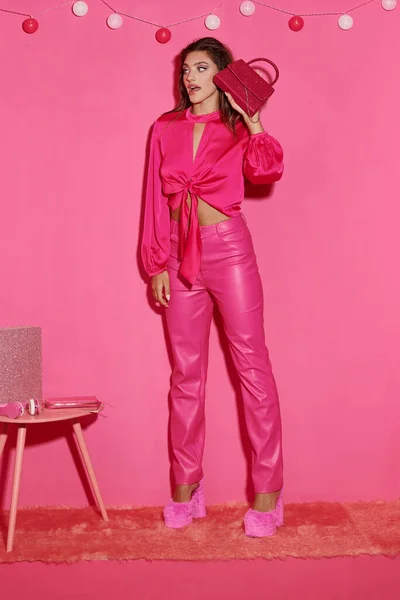 Mujer sorprendida y joven en top de la cosecha y pantalones de color rosa de pie con bolso elegante cerca de la guirnalda de la bola - foto de stock