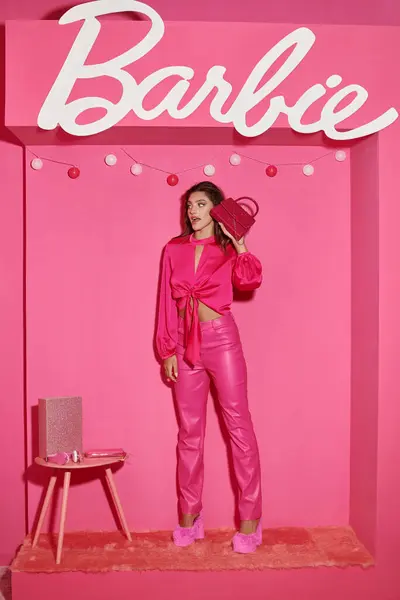 KYIV, UCRANIA - 14 de julio de 2023: sorprendido y muñeca como la mujer en la parte superior de la cosecha y pantalones de color rosa de pie con bolso de mano elegante cerca de signo de Barbie - foto de stock