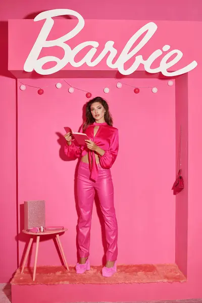 KIEW, UKRAINE - 14. JULI 2023: puppenhafte Frau in bauchfreiem Top und pinkfarbener Hose mit Buch in der Nähe von Barbie-Zeichen und Ballgirlanden — Stockfoto