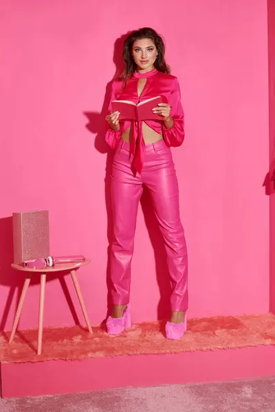 Poupée heureuse comme une femme en haut de culture et pantalon rose debout avec livre et regardant la caméra — Photo de stock