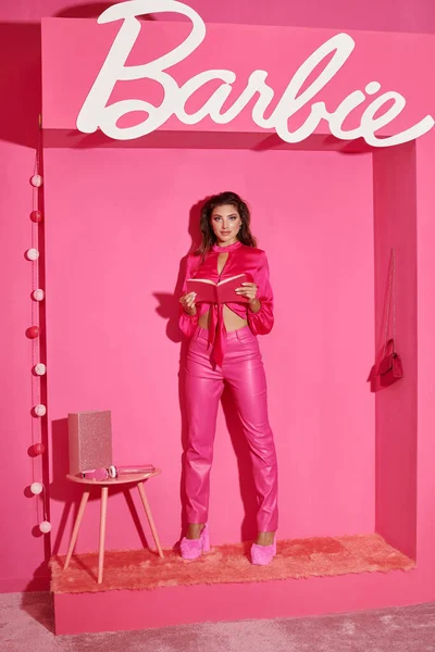 QUIIV, UCRÂNIA - JULHO 14, 2023: boneca bonita como mulher em top de colheita e calças rosa de pé com livro perto de Barbie sinal, glamour — Fotografia de Stock