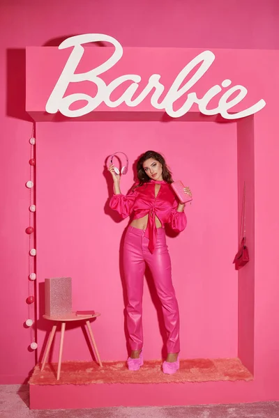 QUIIV, UCRÂNIA - JULHO 14, 2023: boneca bonita como mulher em top de colheita e calças rosa de pé com fones de ouvido perto da assinatura Barbie — Fotografia de Stock