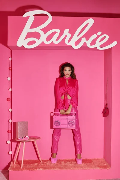 KYIV, UKRAINE - 14 JUILLET 2023 : jeune femme aux cheveux bouclés posant en tenue rose vif et tenant boombox rétro près de signe Barbie — Photo de stock