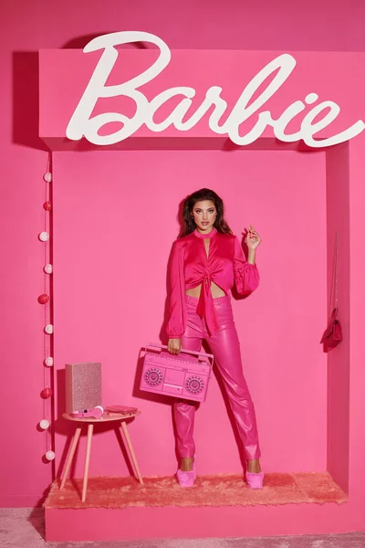 KYIV, UCRANIA - 14 de julio de 2023: bonita joven con el pelo rizado de pie en traje rosa y la celebración de la caja de resonancia cerca de Barbie signo - foto de stock