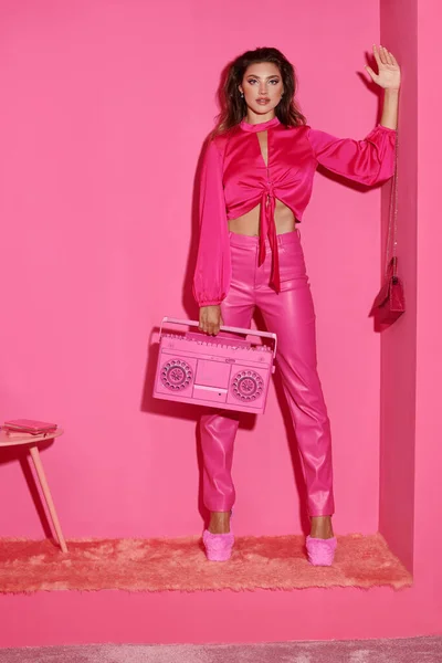 Hermosa mujer joven en traje vibrante sosteniendo boombox retro y agitando la mano sobre fondo rosa - foto de stock