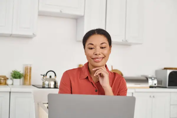 Trabajo remoto de joven nutricionista afroamericana alegre usando el ordenador portátil en su cocina, mujer - foto de stock