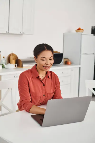 Cheerful young african american nutritionist using laptop while working remotely from her kitchen — Stock Photo