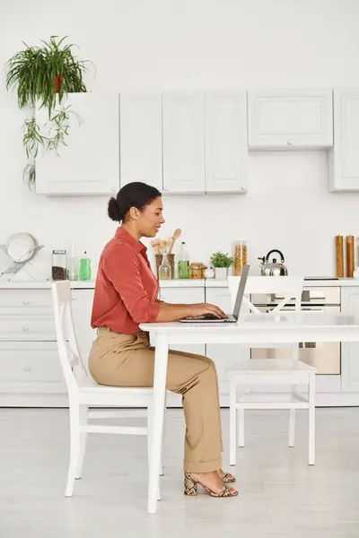 Nutricionista afroamericana satisfecha usando el ordenador portátil mientras trabaja de forma remota desde su cocina moderna - foto de stock