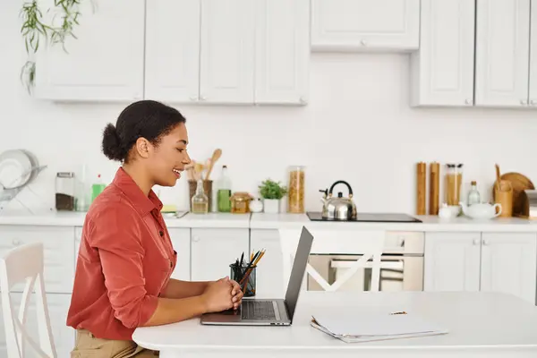 Allegra donna afro-americana che utilizza il computer portatile mentre lavora dalla sua cucina moderna, nutrizionista — Foto stock