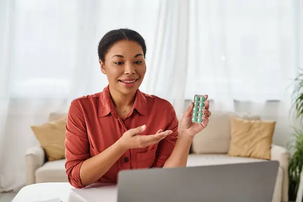 Dietista femenina alegre mostrando medicamentos durante una consulta en línea en el ordenador portátil de la cocina - foto de stock