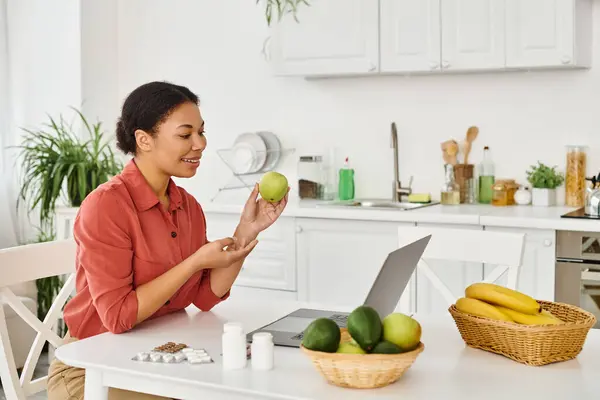 Nutricionista afroamericano feliz sosteniendo manzana mientras que da consejos de dieta en el ordenador portátil en la cocina - foto de stock