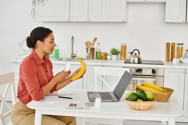 Nutricionista afroamericano feliz sosteniendo plátano y dando consejos de dieta en el ordenador portátil en la cocina - foto de stock