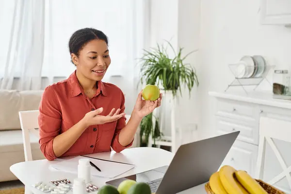 Nutricionista afroamericano feliz sosteniendo manzana verde y dando consejos de dieta en el ordenador portátil en la cocina - foto de stock