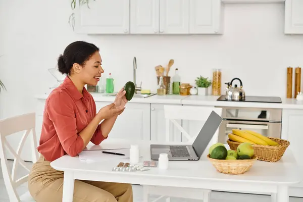 Felice nutrizionista afroamericano che tiene avocado maturo e dà consigli dietetici sul laptop in cucina — Foto stock