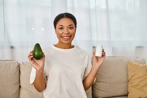 Happy african american nutritionist compares supplements with fresh avocado for a healthy diet — Stock Photo