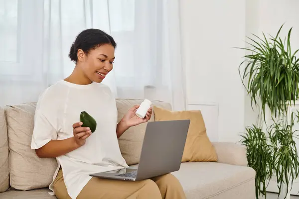 Happy african american nutritionist with supplements and avocado giving dietary advice via laptop — Stock Photo