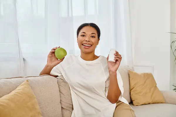 Feliz dietista afro-americano segurando maçã verde e suplementos e sorrindo na sala de estar — Fotografia de Stock