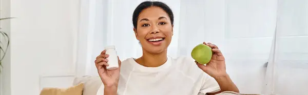 Dietista afroamericano sosteniendo manzana y suplementos y sonriendo en la sala de estar, pancarta - foto de stock