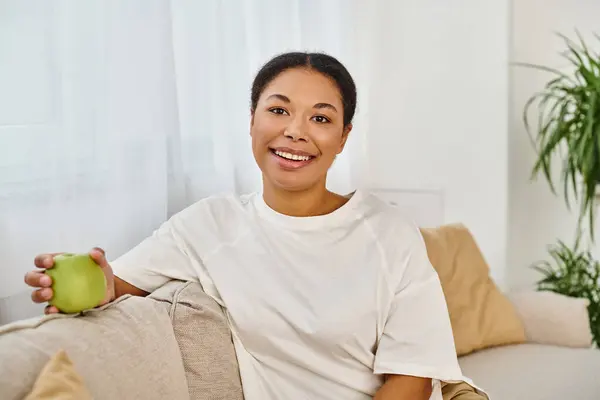 Radiant african american dietitian holding fresh green apple, relaxing on the living room sofa — Stock Photo