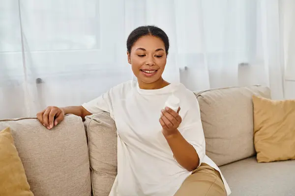 Feliz dietista afro-americano olhando para garrafa com suplementos, relaxando no sofá na sala de estar — Fotografia de Stock