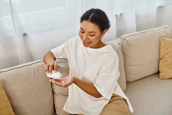 Happy african american dietitian pouring supplements into hand and relaxing on sofa in living room — Stock Photo