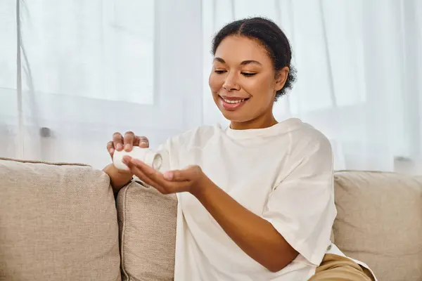 Sonriente dietista afroamericano verter suplementos en la mano y relajarse en el sofá en la sala de estar - foto de stock