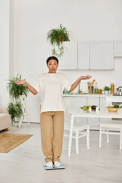 Confused black woman in casual attire standing on scale in kitchen, weight management routine — Stock Photo