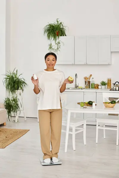 Glückliche schwarze Frau in legerer Kleidung, die in der Küche auf der Waage steht und Nahrungsergänzungsmittel und Apfel in der Hand hält — Stockfoto