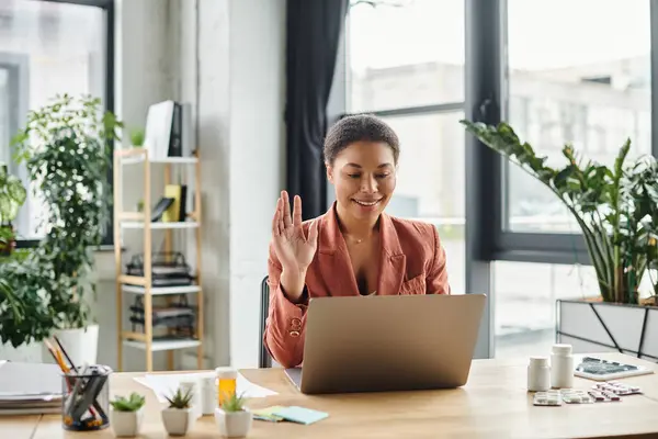 Happy african american nutritionist waving hand at laptop from her plant-filled office space — Stock Photo