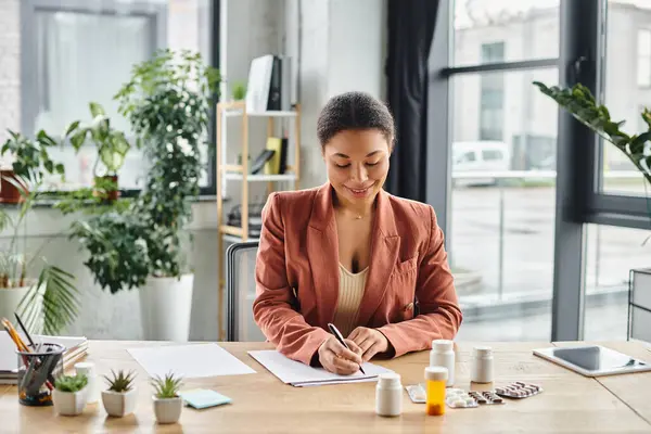 Happy african american nutritionist evaluating medication while writing notes in her office — Stock Photo