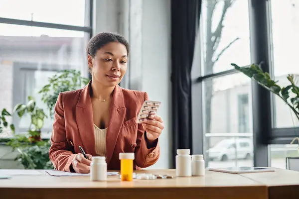 Curly african american nutritionist evaluating medication while writing notes in her office — Stock Photo