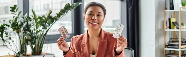 Nutricionista afroamericano alegre mostrando diferentes medicamentos mientras está sentado en la oficina, pancarta - foto de stock