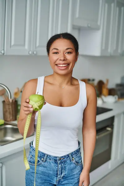 Happy african american woman with measuring tape and apple promoting a healthy diet in kitchen — Stock Photo