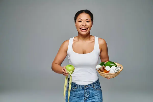 Joyeuse diététiste afro-américaine avec pomme et suppléments mettant en valeur la santé sur fond gris — Photo de stock