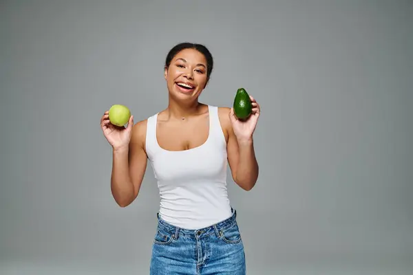 Сияющая африканская американка с яблоком и авокадо, способствующая сбалансированному питанию на сером фоне — стоковое фото