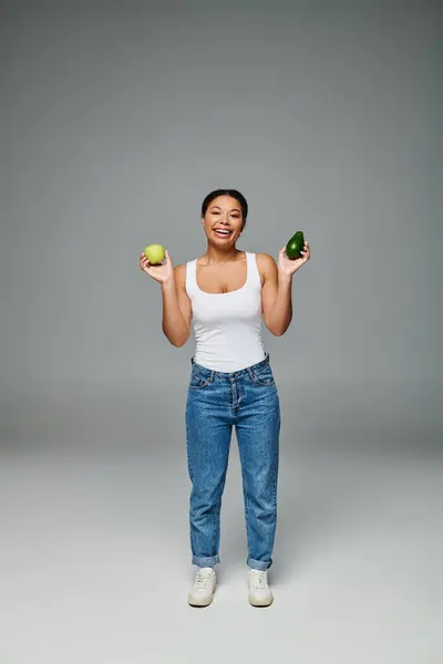 Mujer afroamericana feliz con manzana y aguacate promoviendo una nutrición equilibrada en el fondo gris - foto de stock