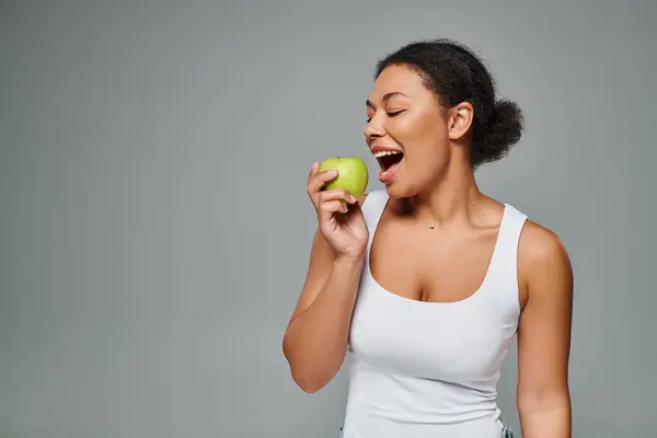 Mujer afroamericana feliz con dientes blancos mordiendo manzana verde sobre fondo gris, alimentación saludable - foto de stock