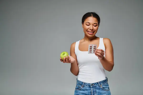 Happy african american woman comparing supplements with green apple grey background, balanced diet — Stock Photo