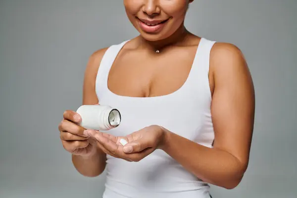Femme afro-américaine cultivée versant des pilules de la bouteille dans la main sur fond gris, plan alimentaire — Photo de stock