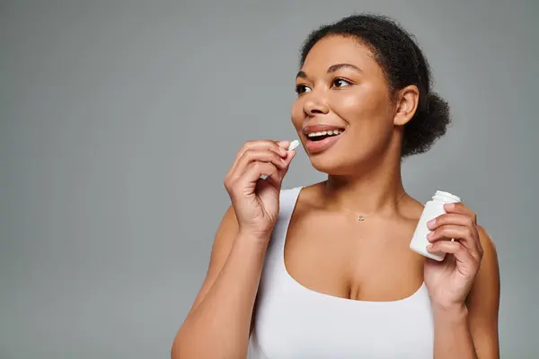 Mujer afroamericana feliz tomando suplementos en forma de píldora sobre fondo gris, plan dietético - foto de stock