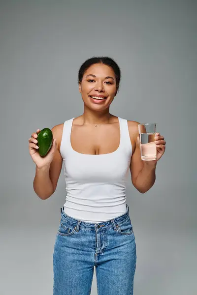 Happy african american nutritionist promoting hydration and healthy eating with avocado and water — Stock Photo