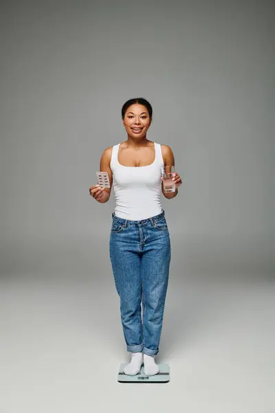 Felice donna afroamericana in possesso di un bicchiere d'acqua e integratori e in piedi su bilance su grigio — Foto stock