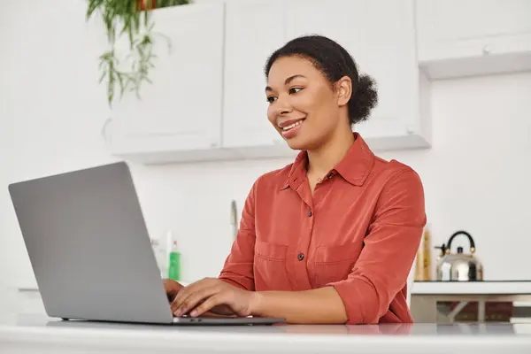 Sonriente dietista afroamericana rizada usando su computadora portátil mientras escribe y trabaja desde casa - foto de stock