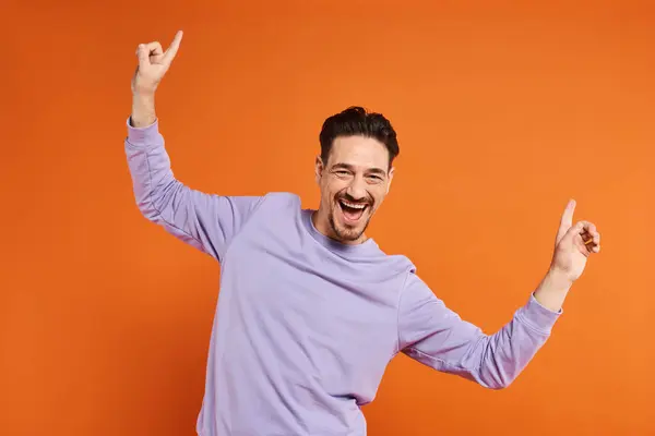 Homem alegre e barbudo em camisola roxa dançando com as mãos no fundo laranja vibrante — Fotografia de Stock